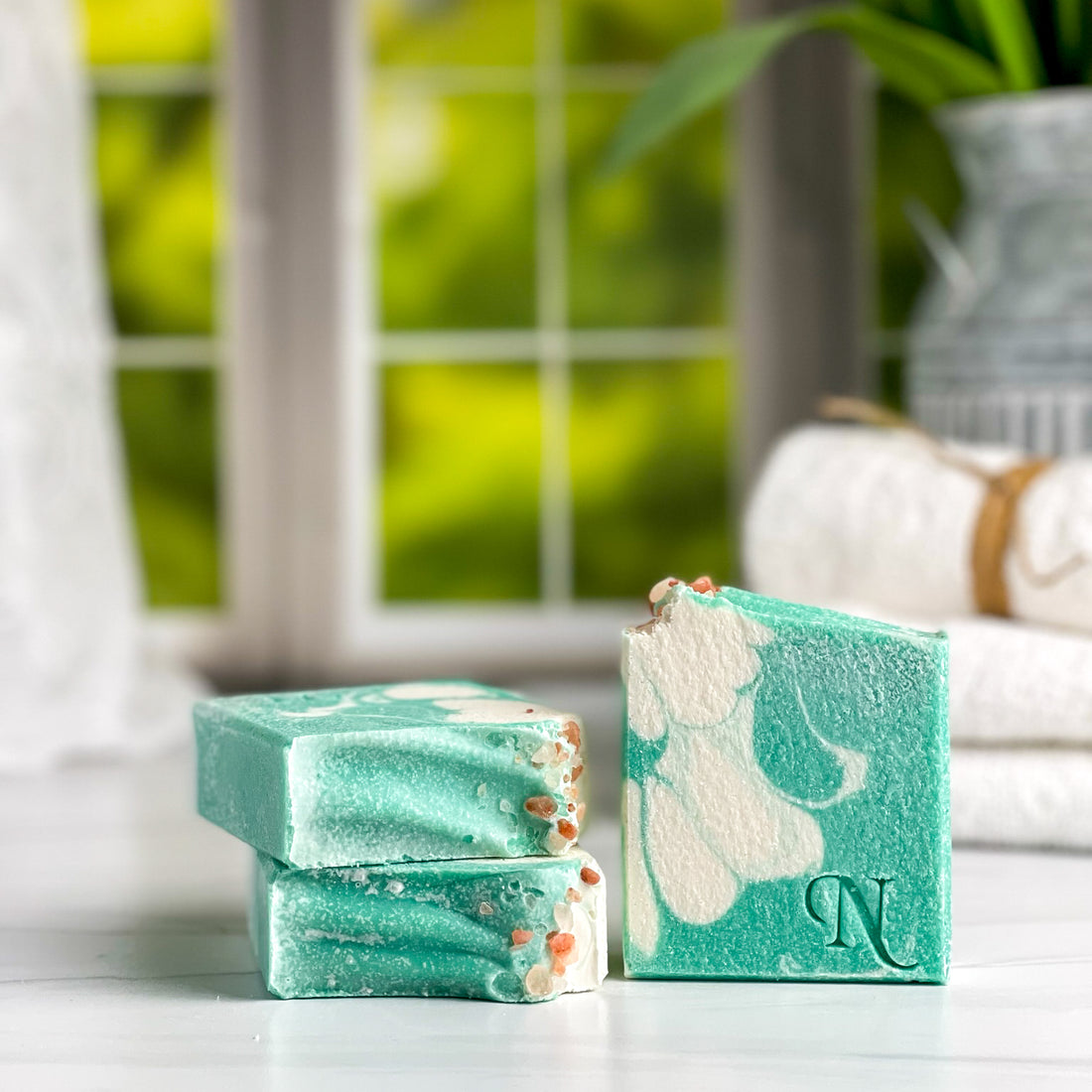 Aura Salt Soap - 3 teal green and white soaps stacked in pile of 2 and one on the side with folded towels in the background